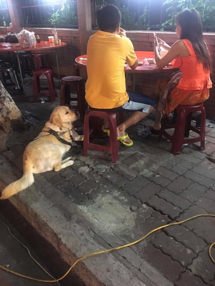 在路邊的騎樓上，一張紅色的大圓桌，冠霖和家人比鄰坐在椅子上靠著桌子，ED趴在冠霖的腳邊